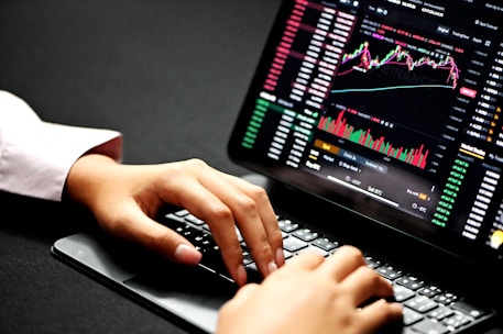 Hands typing on a laptop keyboard with a screen displaying financial graphs and stock market data. Multiple charts and numbers are visible, indicating online trading or financial analysis.