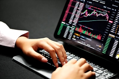 Hands typing on a laptop keyboard with a screen displaying financial graphs and stock market data. Multiple charts and numbers are visible, indicating online trading or financial analysis.
