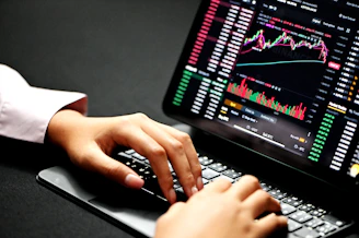Close-up of hands typing on a laptop with financial charts on the screen, set against a clean blue and white workspace.