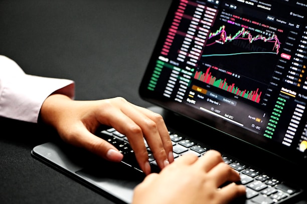 Hands typing on a laptop keyboard with a screen displaying financial graphs and stock market data. Multiple charts and numbers are visible, indicating online trading or financial analysis.