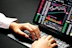 Hands typing on a laptop keyboard with a screen displaying financial graphs and stock market data. Multiple charts and numbers are visible, indicating online trading or financial analysis.