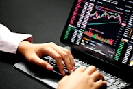 Close-up of hands typing on a laptop with financial charts on the screen.