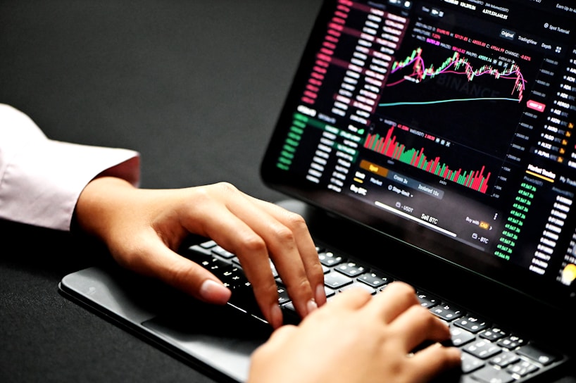 Hands typing on a laptop keyboard with a screen displaying financial graphs and stock market data. Multiple charts and numbers are visible, indicating online trading or financial analysis.