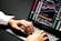 Hands typing on a laptop keyboard with a screen displaying financial graphs and stock market data. Multiple charts and numbers are visible, indicating online trading or financial analysis.