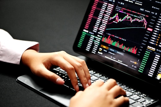 Hands typing on a laptop keyboard with a screen displaying financial graphs and stock market data. Multiple charts and numbers are visible, indicating online trading or financial analysis.