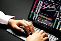 Hands typing on a laptop keyboard with a screen displaying financial graphs and stock market data. Multiple charts and numbers are visible, indicating online trading or financial analysis.