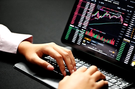 Hands typing on a laptop keyboard with a screen displaying financial graphs and stock market data. Multiple charts and numbers are visible, indicating online trading or financial analysis.
