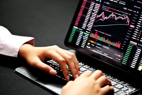 Hands typing on a laptop keyboard with a screen displaying financial graphs and stock market data. Multiple charts and numbers are visible, indicating online trading or financial analysis.