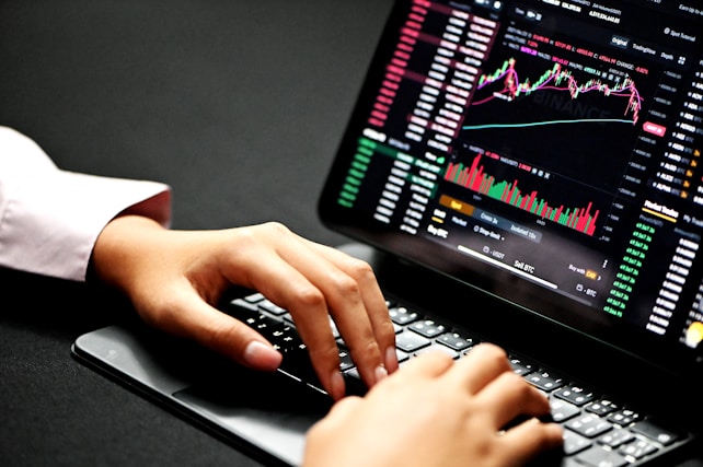 Hands typing on a laptop keyboard with a screen displaying financial graphs and stock market data. Multiple charts and numbers are visible, indicating online trading or financial analysis.