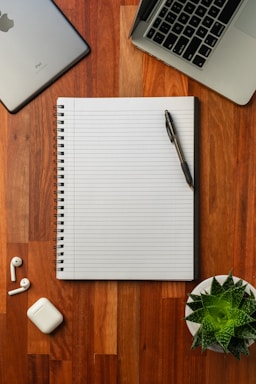 A cozy wooden desk with a notebook, pen, and a small pot of fresh ginseng roots beside a laptop.