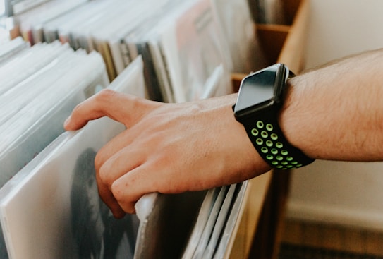 A hand wearing a smartwatch with a green and black band is reaching to select a record from a collection. The records are stacked vertically in sleeves, and the scene suggests a personal or retail environment with wooden furniture and soft lighting.