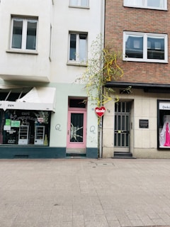 A street view featuring a section of a row of buildings, showcasing different architectural styles with a mix of white and brick facades. A storefront with a pink door is visible, alongside neighboring businesses. A small tree with sparse yellow-green leaves is situated in front, and a heart-shaped sign with the word 'Nicole' hangs prominently. Windows of varying designs are seen, and the sidewalk is wide and paved.