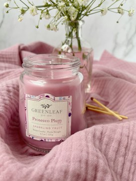 A pink candle labeled 'Greenleaf Prosecco Plum Sparkling Fruit' is surrounded by soft pink fabric. In the background, a glass vase holds delicate white flowers against a light, textured backdrop.