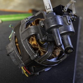 A close-up view of a mechanical device featuring metallic components with visible screws and a coiled copper wire. Multiple wires of different colors, including green, yellow, and red, are attached to the device. The surface is slightly textured, and the surrounding area appears to be part of a workshop or industrial setting.