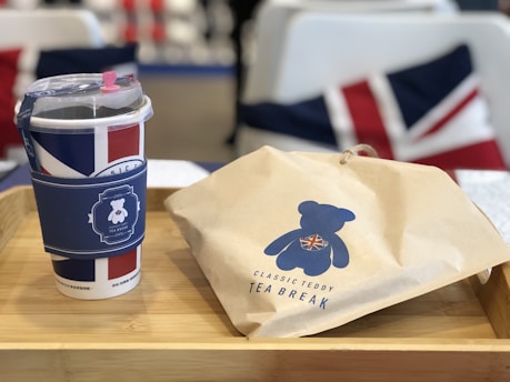 A wooden tray holds a cup and a paper bag. The cup features designs in colors resembling the Union Jack and a decorative sleeve with a teddy bear motif. The paper bag also has a teddy bear design, labeled 'Classic Teddy Tea Break.' In the background, there are cushions with similar patterns.