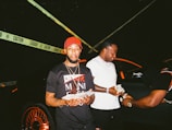 Three men are standing next to a black car with orange highlights. One wears a black and red cap and a shirt with a cigar in his mouth, holding money. Another wears a white shirt and chain, also holding money, while a third man dressed in black and orange bends down slightly, engaging with the money. Yellow caution tape is stretched behind them.