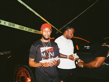 Three men are standing next to a black car with orange highlights. One wears a black and red cap and a shirt with a cigar in his mouth, holding money. Another wears a white shirt and chain, also holding money, while a third man dressed in black and orange bends down slightly, engaging with the money. Yellow caution tape is stretched behind them.