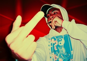 A person wearing a white hoodie with a colorful design prominently featuring a yin-yang symbol and floral elements. They are holding a cigar or similar object near their mouth, with the other hand extended forward, displaying the middle finger. The background is vibrant red, enhancing the overall intensity of the image.