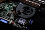 A close-up view of the internal components of a computer, highlighting the circuit board and cooling fan. Various electronic components such as capacitors, connectors, and integrated circuits are visible. Colorful wires are connected to the motherboard, and there are QR codes and small printed text on some parts.