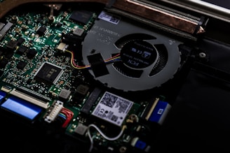 A close-up view of the internal components of a computer, highlighting the circuit board and cooling fan. Various electronic components such as capacitors, connectors, and integrated circuits are visible. Colorful wires are connected to the motherboard, and there are QR codes and small printed text on some parts.