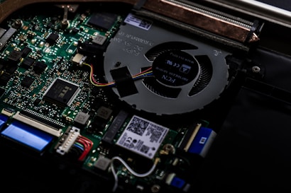 A close-up view of the internal components of a computer, highlighting the circuit board and cooling fan. Various electronic components such as capacitors, connectors, and integrated circuits are visible. Colorful wires are connected to the motherboard, and there are QR codes and small printed text on some parts.