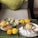 A beautifully arranged platter of Indian snacks and appetizers is displayed, featuring samosas, dhokla, and chutneys. Accompanying these dishes are decorative marigold flowers, enhancing the presentation. The setting is cozy, with a plush cushion in the background.