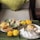 A beautifully arranged platter of Indian snacks and appetizers is displayed, featuring samosas, dhokla, and chutneys. Accompanying these dishes are decorative marigold flowers, enhancing the presentation. The setting is cozy, with a plush cushion in the background.
