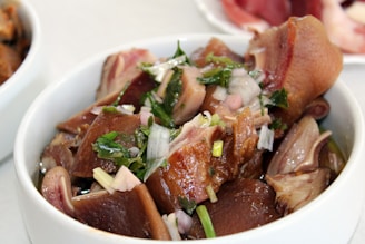 A white bowl filled with sliced pieces of cooked meat, garnished with chopped onions and fresh green herbs. The dish appears seasoned, showcasing glossy textures with hints of juice or broth.