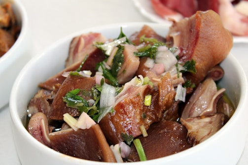 A white bowl filled with sliced pieces of cooked meat, garnished with chopped onions and fresh green herbs. The dish appears seasoned, showcasing glossy textures with hints of juice or broth.