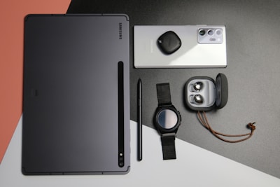 A flat lay of various Samsung electronic devices and accessories, including a tablet, a smartphone with a tag attached, a pen, a smartwatch with a black strap, and a pair of wireless earbuds in an open charging case, all arranged on a multicolored background with shades of black, gray, white, and peach.