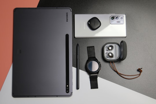 A flat lay of various Samsung electronic devices and accessories, including a tablet, a smartphone with a tag attached, a pen, a smartwatch with a black strap, and a pair of wireless earbuds in an open charging case, all arranged on a multicolored background with shades of black, gray, white, and peach.