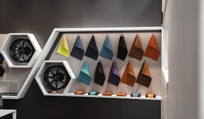 A modern wall display features several fabric samples in triangular shapes, each attached at the top corners. The samples include various colors such as green, blue, brown, and black. To the left, black alloy wheels are mounted inside hexagonal white frames. The setting is contemporary, likely within a showroom or design studio, highlighting materials and automotive parts.
