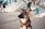 A German Shepherd dog is sitting attentively on a cobblestone street with a blurred background that includes people walking and a set of stairs leading to a building. The dog has a focused expression, and the warm sunlight casts a soft glow on its fur.