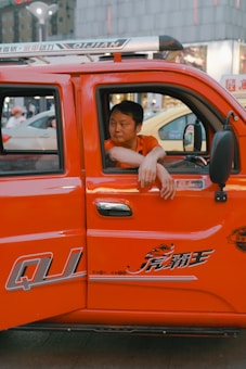 A person is sitting in the driver's seat of an orange vehicle, with their arm resting on the window. The vehicle is a utility truck, highlighted by its bright orange color and distinct decals. The setting appears urban, with buildings and street lights visible in the background.