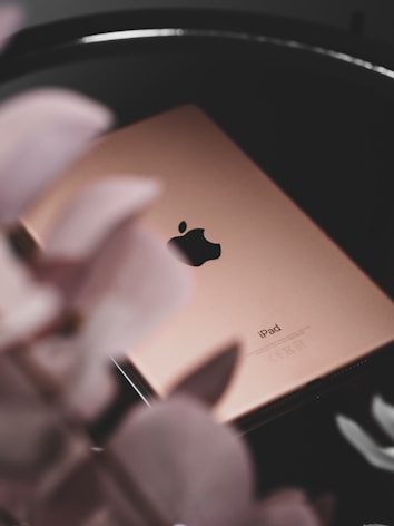 A gold-colored iPad with the Apple logo is placed on a dark reflective surface. Blurred petals in the foreground add a soft, elegant touch to the composition.