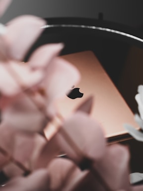 A gold-colored Apple laptop partially obscured by soft focus pink flowers, creating a serene and elegant composition. The background is blurred, highlighting the sleek design of the laptop.