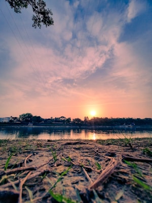 The image captures a serene sunset over a calm river, with a sandy shore in the foreground. Sparse grass and twigs are scattered across the sand. The sun is setting on the horizon, giving the sky a blend of warm orange and cool blue hues. Reflections of the trees and sky are visible in the river, and there are power lines faintly running across the sky. A few buildings are visible among the trees along the riverbank.