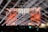 A large concert stage with bright spotlights illuminating a crowd. The backdrop is adorned with vibrant orange, red, and gold patterns, resembling a futuristic or fantasy architecture. Beams of white light crisscross over the audience, creating a dynamic and energetic atmosphere.