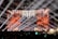 A large concert stage with bright spotlights illuminating a crowd. The backdrop is adorned with vibrant orange, red, and gold patterns, resembling a futuristic or fantasy architecture. Beams of white light crisscross over the audience, creating a dynamic and energetic atmosphere.
