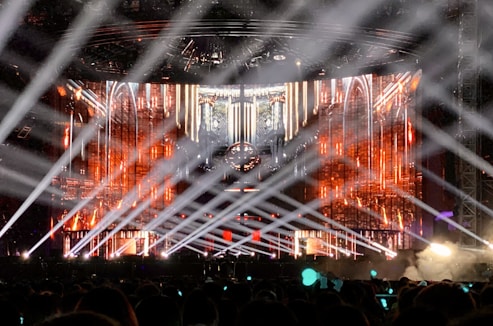A large concert stage with bright spotlights illuminating a crowd. The backdrop is adorned with vibrant orange, red, and gold patterns, resembling a futuristic or fantasy architecture. Beams of white light crisscross over the audience, creating a dynamic and energetic atmosphere.