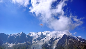 A picturesque view of snow-capped mountains with a bright blue sky and scattered clouds. A single paraglider is flying high above the mountain peaks, creating a sense of adventure and freedom.