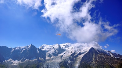 A picturesque view of snow-capped mountains with a bright blue sky and scattered clouds. A single paraglider is flying high above the mountain peaks, creating a sense of adventure and freedom.