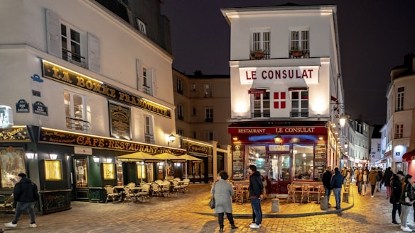 A charming street scene features a historic French cafe and restaurant with outdoor seating. Warm lights illuminate the cobblestone street, while several people walk around or sit at tables. The buildings are adorned with classic French signage and windows framed by shutters.
