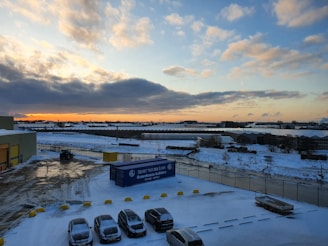 A snow-covered industrial area during sunset, with a cloudy sky. Several parked cars are visible in the foreground, alongside some industrial buildings. There's a blue shipping container labeled 'Greenhouse builders' near one building. The landscape extends to greenhouse structures in the background.