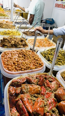 A display of various pickled vegetables and condiments in large, clear containers. Each container is filled with different types of pickles, including ones with bright red chilies, green chilies, and chickpeas. The containers are neatly arranged on a table, and several metal spoons are placed in them for serving. In the background, a person is visible, possibly a vendor, attending to the stall.