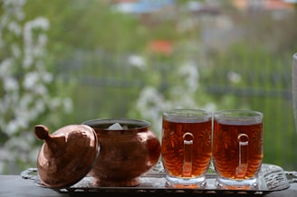 A copper sugar bowl with a lid is placed alongside two glass mugs filled with tea, set on an ornate metal tray. The background features a blurred green outdoor scene.