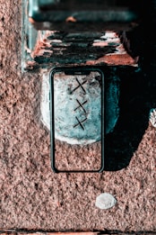 A smartphone case placed on a surface, possibly concrete or stone, with three crossed black marks (XXX) on the surface inside the case frame. The surface appears to be weathered, with texture and some paint remnants.
