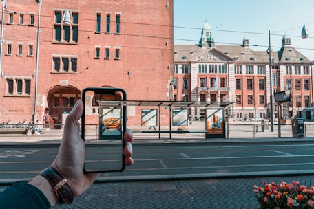 A hand holding a smartphone captures a city street scene where a historic building with a detailed facade is visible. The phone screen aligns seamlessly with the building's background. A tram station and tall red brick architecture are prominent features, and there are advertisements visible on the station platforms. Tulips appear in the foreground.