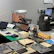 A workbench is cluttered with technology repair tools and disassembled electronic devices. Multiple laptops are open, and an overhead lamp provides focused lighting on the workspace. A backpack sits at the back, amidst a disarray of tools such as screwdrivers, a utility knife, and an organized box of small components. Various pieces of electronic hardware and circuit boards are spread out on the table.