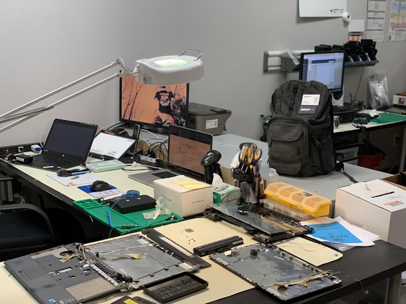 A workbench is cluttered with technology repair tools and disassembled electronic devices. Multiple laptops are open, and an overhead lamp provides focused lighting on the workspace. A backpack sits at the back, amidst a disarray of tools such as screwdrivers, a utility knife, and an organized box of small components. Various pieces of electronic hardware and circuit boards are spread out on the table.